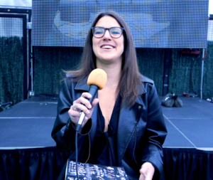 Sam Sumpter, a woman with brown hair and glasses, holds a microphone and smiles.