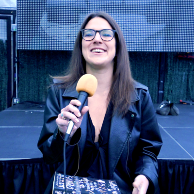 Sam Sumpter, a woman with brown hair and glasses, holds a microphone and smiles.