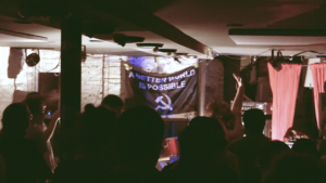 a still from friendship quest fest video depicting a person singing in a crowded room, in front of a banner that says A BETTER WORLD IS POSSIBLE