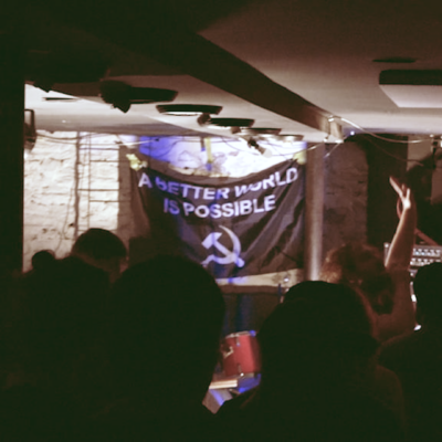 a still from friendship quest fest video depicting a person singing in a crowded room, in front of a banner that says A BETTER WORLD IS POSSIBLE