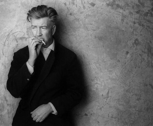 Black and white photo of David Lynch, smoking against a wall. 